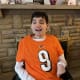 A smiling young man with CP, wearing an orange number nine sports shirt, sits in his wheelchair in front of a stone mantelpiece